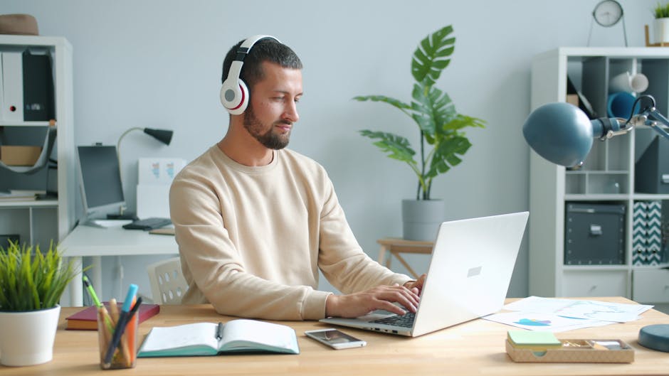 Adult man with headphones working on laptop in home office setting.