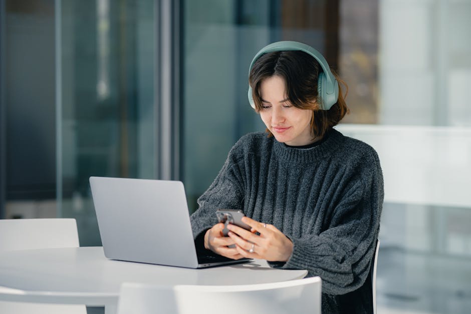 A young woman wearing headphones uses a laptop and smartphone indoors at a bright table.