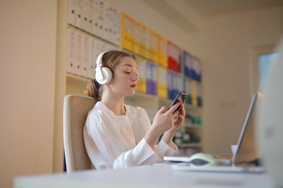 Young woman in white shirt using smartphone and headphones at her office desk.